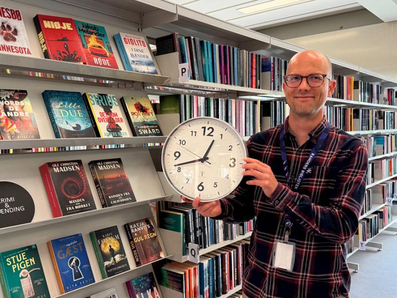 Mads Einar Nielsen holder et væg-ur i biblioteksrummet.