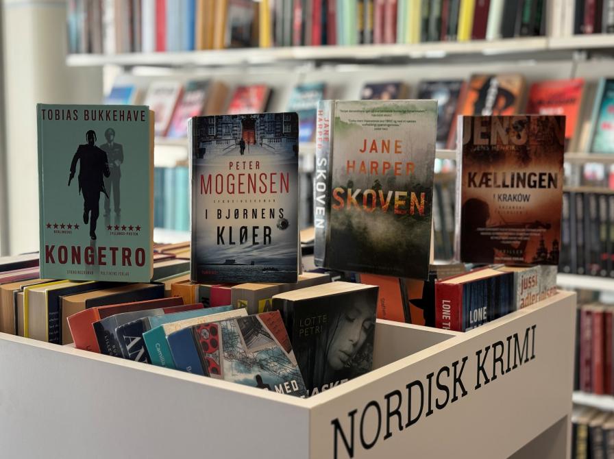 Fire bøger stillet på højkant på en krybbe med bøger med bogreoler i baggrunden